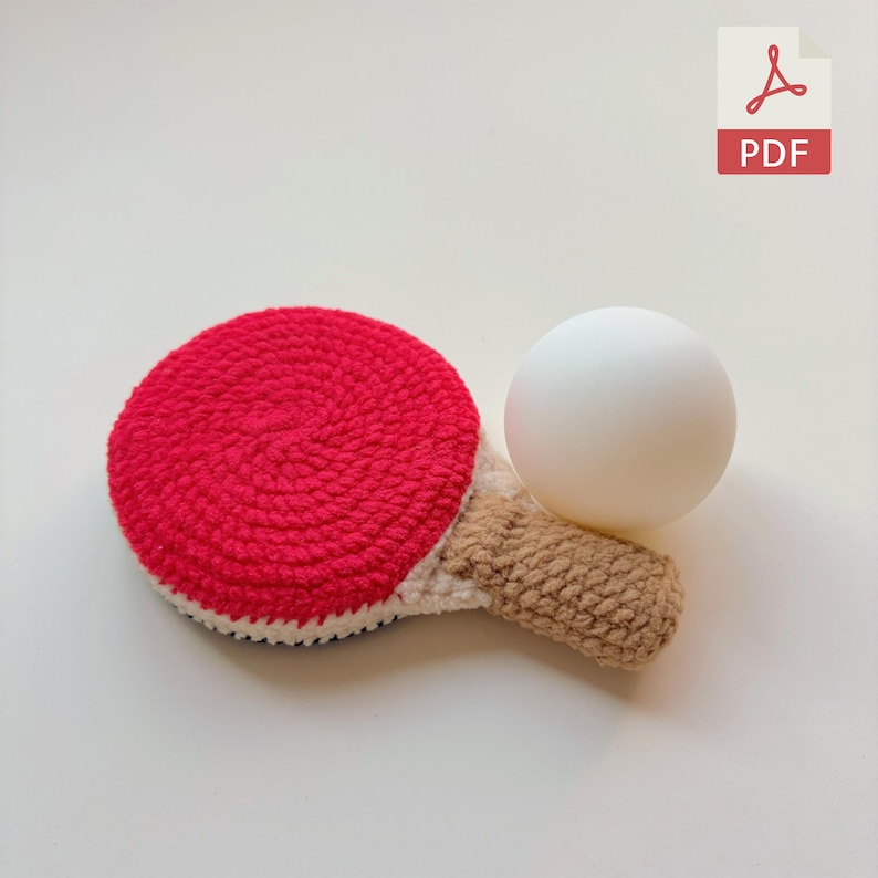 May include: A handmade crochet ping pong paddle with a red head, white and black trim, and a tan handle. A white ping pong ball rests on the paddle. The image includes a PDF icon.