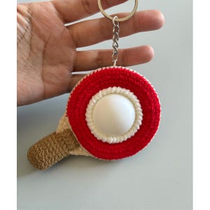 May include: A handmade keychain featuring a red and white crocheted circle with a white center, resembling a paddleball. The handle is crocheted in a tan color. A silver chain and keyring are attached.