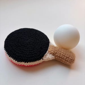 May include: A handmade crochet ping pong paddle with a black playing surface, a white and pink edge, and a tan handle. A white ping pong ball rests beside the paddle. The items are made of yarn.