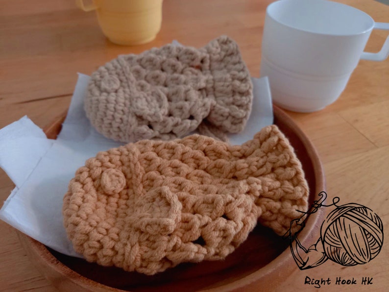 May include: Two crocheted tan and brown  fish-shaped  coasters on a wooden surface. The coasters are made with a textured stitch pattern.