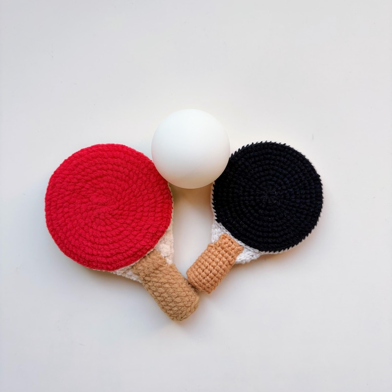 May include: A crochet ping pong set with a red paddle, a black paddle, and a white ball. The paddles have tan handles and white and tan accents. The set is displayed on a white background.