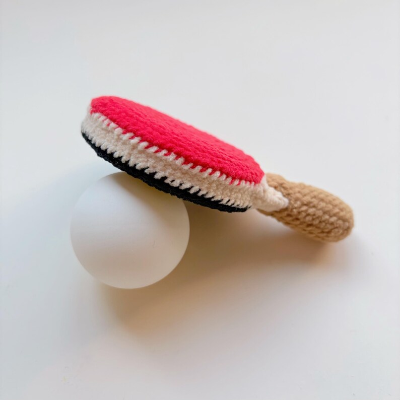 May include: A handmade crochet ping pong paddle with a red, white, and black paddle head and a tan handle. The paddle rests on a white ball, suggesting a game of table tennis. The craft item is likely for a child.