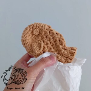 May include: A crocheted tan-colored taiyaki, a Japanese fish-shaped pastry, is held in a hand and wrapped in white tissue paper.