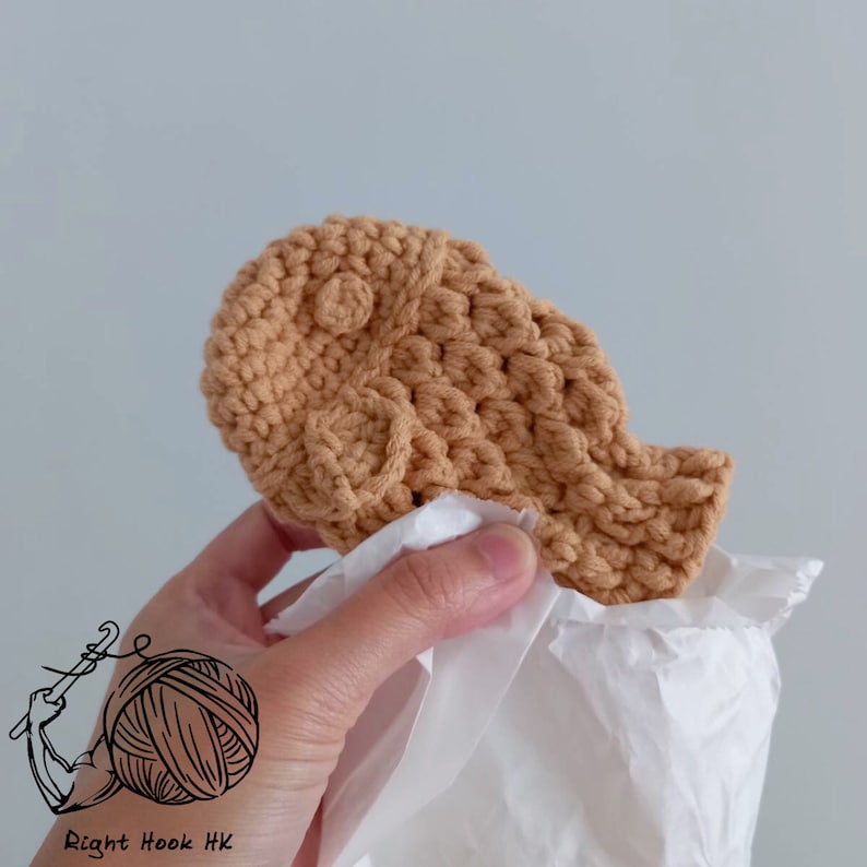May include: A crocheted tan-colored taiyaki, a Japanese fish-shaped pastry, is held in a hand and wrapped in white tissue paper.