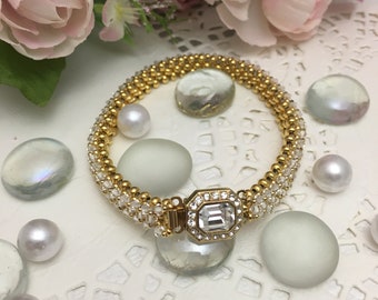 May include: A gold bracelet with a clear crystal centerpiece and a row of clear beads surrounding it. The bracelet is on a white surface with white and clear glass beads.