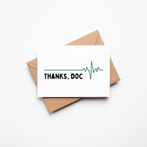Puede incluir: Una tarjeta de felicitación blanca con el texto "THANKS, DOC" en negro, acompañada de un diseño de línea de latido cardíaco verde. La tarjeta está colocada sobre un sobre marrón, sobre un fondo blanco.
