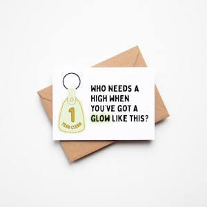 May include: A greeting card with a brown envelope. The card has the text "WHO NEEDS A HIGH WHEN YOU'VE GOT A GLOW LIKE THIS?" and a keychain that says "1 YEAR CLEAN". The keychain is light yellow with a black ring.