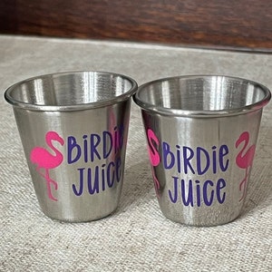May include: Two stainless steel shot glasses with a pink flamingo and the words "Birdie Juice" in purple. The shot glasses are cylindrical with a rolled rim and are sitting on a neutral-colored surface.