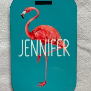 May include: A teal blue luggage tag featuring a pink flamingo illustration and the name "Jennifer" in white text.