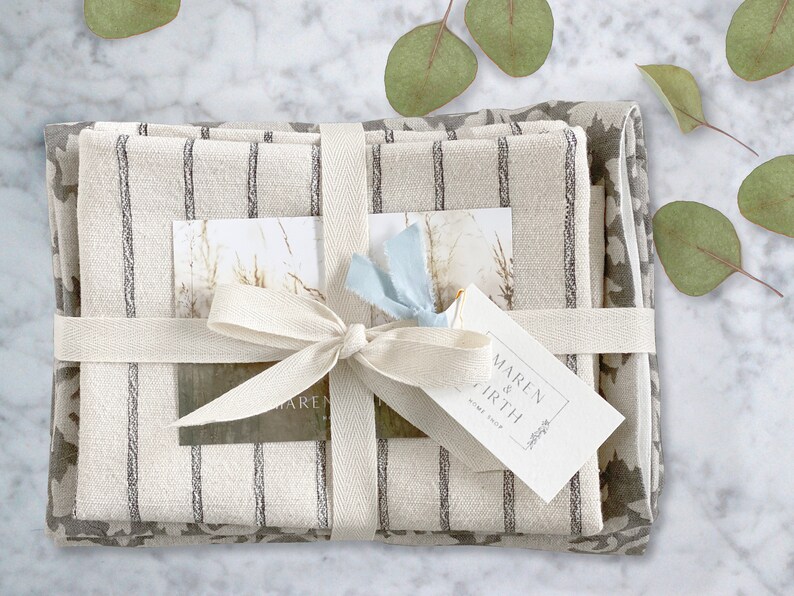 May include: A gift set of three linen kitchen towels, wrapped in a white ribbon with a bow. The towels are striped in grey and white and feature a floral pattern. The gift tag reads "MAREN & EARTH HOME SHOP".