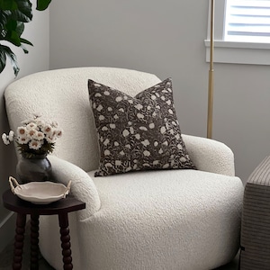 K&ouml;nnte beinhalten: Ein wei&szlig;er, flauschiger Sessel mit einem braunen Kissen mit Blumenmuster. Der Sessel steht in einem Wohnzimmer mit einem Beistelltisch und einer Stehlampe.