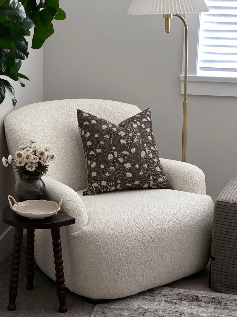 K&ouml;nnte beinhalten: Ein wei&szlig;er, flauschiger Sessel mit einem braunen Kissen mit Blumenmuster. Der Sessel steht in einem Wohnzimmer mit einem Beistelltisch und einer Stehlampe.