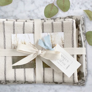 May include: A gift set of three linen kitchen towels, wrapped in a white ribbon with a bow. The towels are striped in gray and white and feature a floral pattern. The gift tag reads "MAREN & EARTH HOME SHOP".