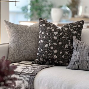 K&ouml;nnte beinhalten: Ein wei&szlig;es Sofa mit drei Kissen. Die Kissen sind mit floralen Mustern in Grau- und Schwarzt&ouml;nen verziert. Eine braune und wei&szlig;e gestreifte Decke liegt auf dem Sofa.