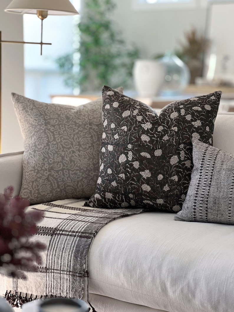 K&ouml;nnte beinhalten: Ein wei&szlig;es Sofa mit drei Kissen. Die Kissen sind mit floralen Mustern in Grau- und Schwarzt&ouml;nen verziert. Eine braune und wei&szlig;e gestreifte Decke liegt auf dem Sofa.