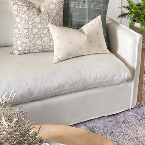 May include: A white linen sofa with three throw pillows. The pillows are patterned with floral designs in beige and brown, a geometric pattern in beige, and a plaid pattern in green. The sofa is sitting on a patterned rug.