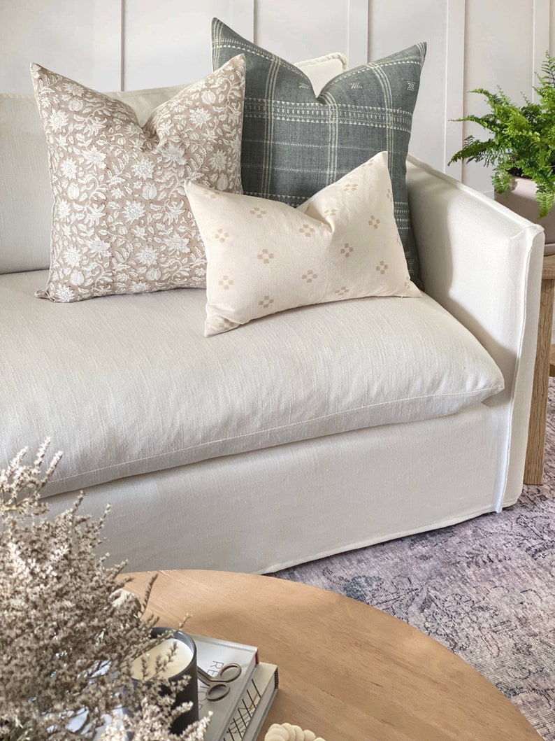 May include: A white linen sofa with three throw pillows. The pillows are patterned with floral designs in beige and brown, a geometric pattern in beige, and a plaid pattern in green. The sofa is sitting on a patterned rug.