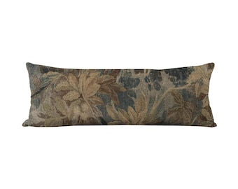 May include: A long, rectangular cushion with a botanical pattern. The cushion features a tapestry-like design with shades of green, brown, and beige, depicting stylized leaves and foliage. The cushion is designed for decorative use on a bed or sofa.