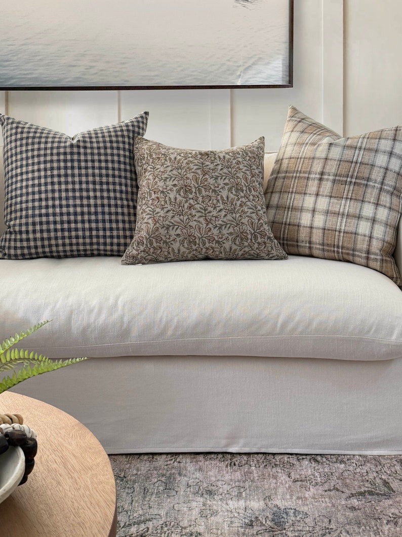 May include: Three throw pillows on a white sofa. The pillows are made of different fabrics and have different patterns. The pillow on the left is a black and white gingham pattern. The middle pillow is a brown floral pattern. The pillow on the right is a brown and white plaid pattern.