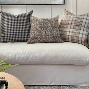 May include: Three throw pillows on a white couch. The pillows are made of different fabrics and patterns. The pillow on the left is a black and white gingham pattern. The middle pillow is a brown floral pattern. The pillow on the right is a brown and white plaid pattern.