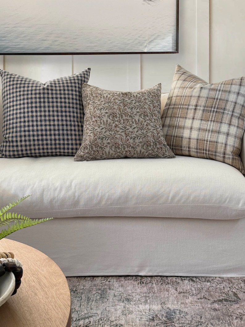 May include: Three throw pillows on a white couch. The pillows are made of different fabrics and patterns. The pillow on the left is a black and white gingham pattern. The middle pillow is a brown floral pattern. The pillow on the right is a brown and white plaid pattern.