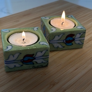 May include: Two green ceramic candle holders with white and blue floral designs. Each holder has a white tea light candle inside.