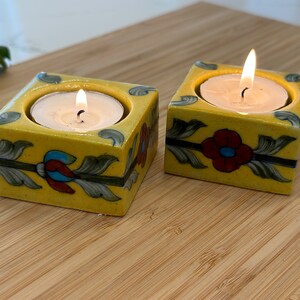 May include: Two yellow ceramic candle holders with floral designs. Each holder has a tealight candle inside, and the flames are lit.