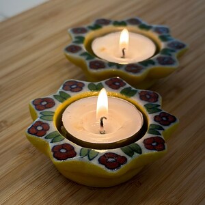 May include: Two yellow ceramic candle holders with a floral design. Each holder has a small white candle inside, and both candles are lit.