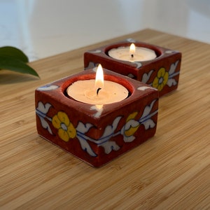 May include: Two terracotta candle holders with floral designs in shades of blue, white, and yellow. Each holder has a small white candle inside, with the flames lit.