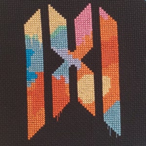 May include: A cross-stitch design featuring a colorful abstract letter "M" against a black background. The letter is composed of various geometric shapes filled with orange, pink, blue, and yellow threads. The design has a handmade, textured appearance.