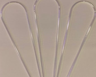 May include: Three clear, U-shaped plastic straws are arranged in a fan shape against a light background. The straws are transparent and have a smooth, curved top and straight sides.