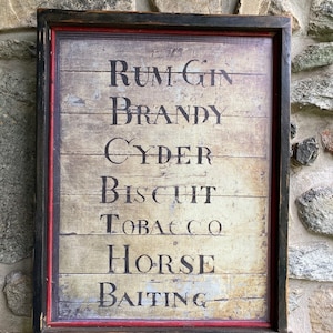 May include: A framed vintage-style sign with a woodgrain background and red trim. The sign lists various goods for sale, including "Rum, Gin, Brandy, Cyder, Biscuit, Tobacco, Horse Baiting."