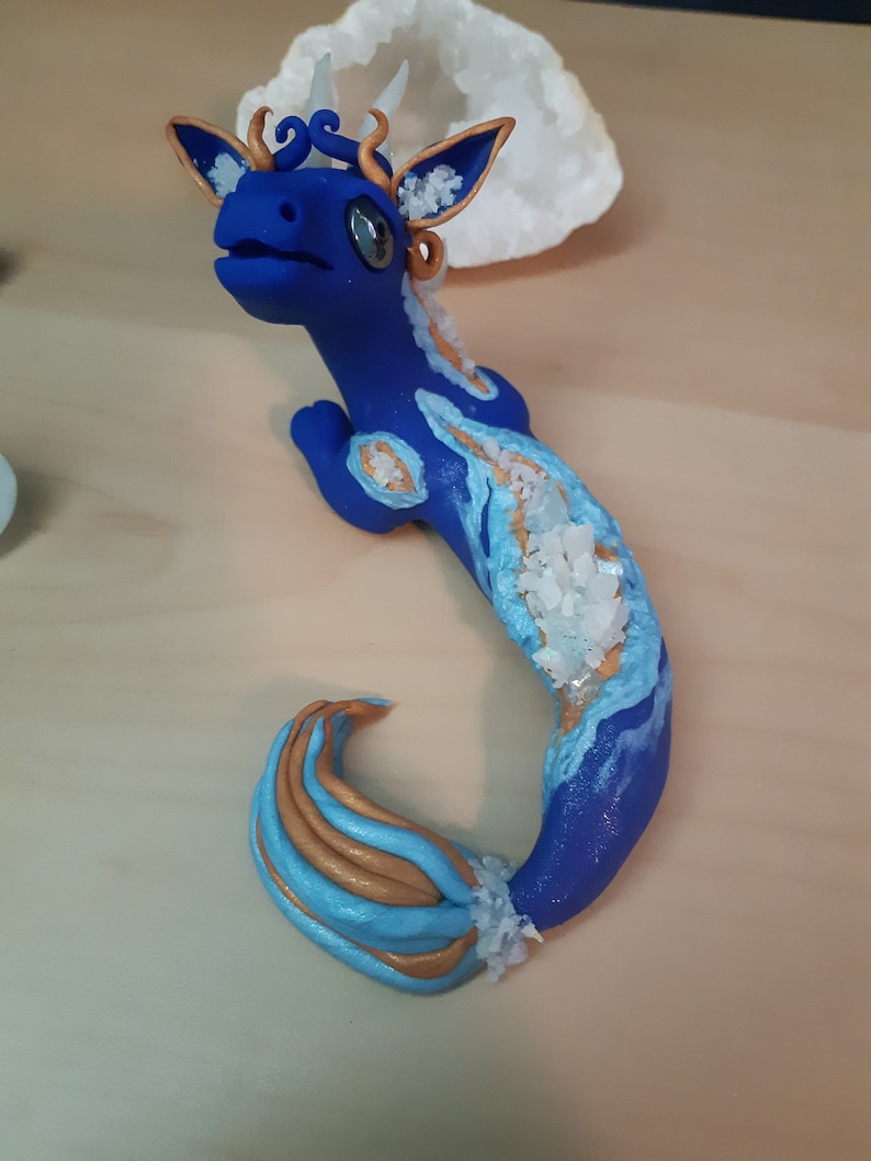 May include: A blue and gold dragon figurine with a crystal-like texture. The dragon has a long tail and a small head with large eyes. The figurine is made of polymer clay and is hand-painted.