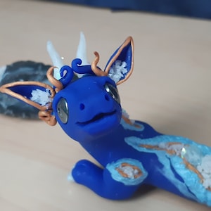 May include: A blue and white dragon figurine with gold accents. The dragon has white horns and a blue body with white and gold details. The dragon is lying on its belly with its head raised.