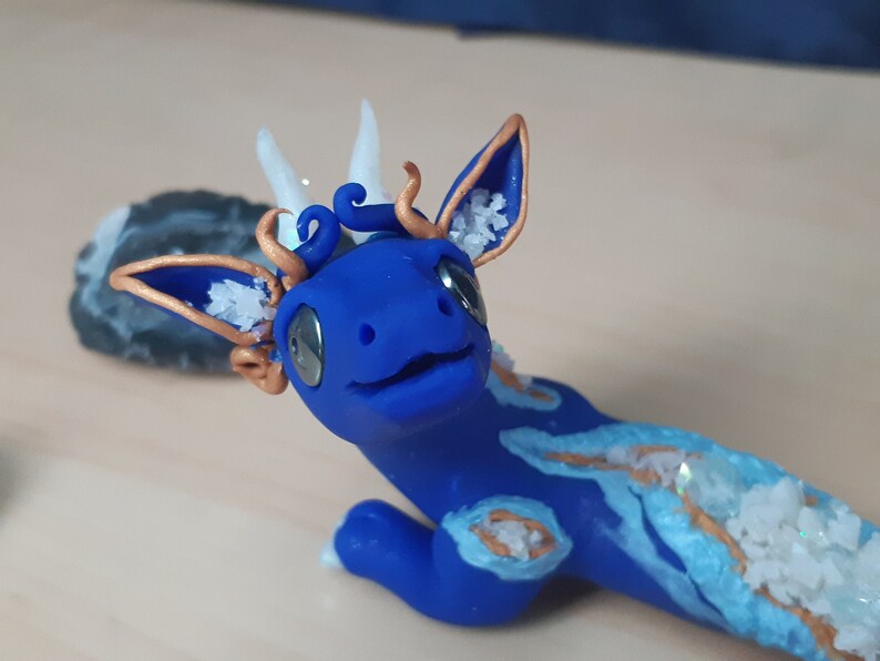 May include: A blue and white dragon figurine with gold accents. The dragon has white horns and a blue body with white and gold details. The dragon is lying on its belly with its head raised.
