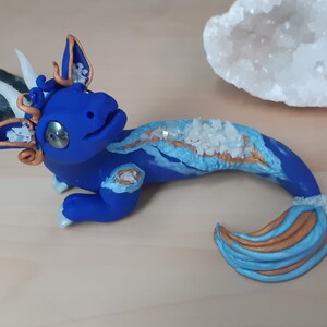 May include: A blue and gold dragon figurine with white crystal accents. The dragon has a curled tail and a whimsical expression.
