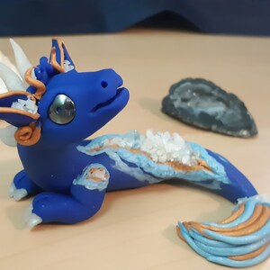 May include: A blue and gold dragon figurine with white crystal accents. The dragon is posed with its head tilted up and its tail curled behind it.