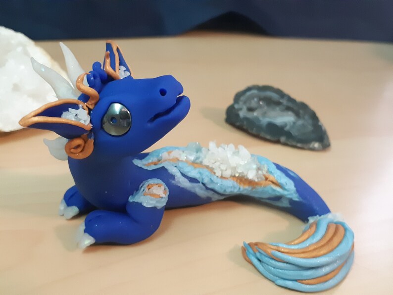May include: A blue and gold dragon figurine with white crystal accents. The dragon is posed with its head tilted up and its tail curled behind it.