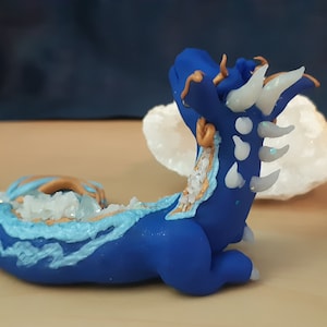 May include: A blue and white ceramic dragon figurine with gold accents. The dragon is curled up in a sleeping position with its head resting on its tail. The dragon is decorated with white and blue crystals.