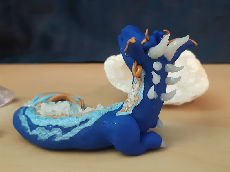 May include: A blue and white ceramic dragon figurine with gold accents. The dragon is curled up in a sleeping position with its head resting on its tail. The dragon is decorated with white and blue crystals.