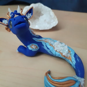 May include: A blue and gold dragon figurine with white crystal accents. The dragon has a long tail and is lying on its belly.
