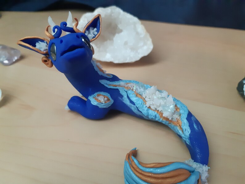 May include: A blue and gold dragon figurine with white crystal accents. The dragon has a long tail and is lying on its belly.