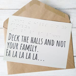 May include: A white greeting card with the text "Deck the Halls and Not Your Family. Fa La La La La..." in black font, with braille dots above the text, inside a brown envelope, on a white wooden surface.