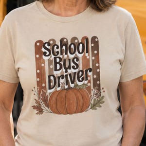 May include: A beige short-sleeve t-shirt featuring a graphic print with the text School Bus Driver in a dark brown font. The design includes a large orange pumpkin, autumn leaves, and a background of vertical brown and white polka-dot stripes.
