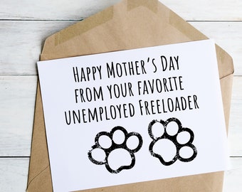 May include: A white Mother's Day card with black text reading "HAPPY MOTHER'S DAY FROM YOUR FAVORITE UNEMPLOYED FREELOADER" and two black paw prints. The card is in a brown envelope, on a white wooden surface.