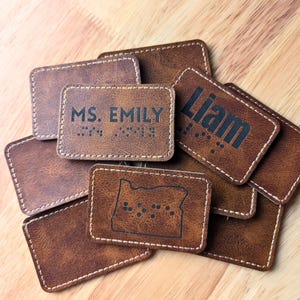 May include: Several brown leather name tags with white stitching are displayed. Some tags have names such as "Liam" and "Ms. Emily" in black lettering, with braille dots. One tag features the outline of Oregon with braille.