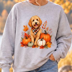 May include: A light gray crewneck sweatshirt featuring a graphic print of a golden-colored dog wearing a plaid bandana, surrounded by orange and white pumpkins and vibrant autumn leaves in shades of red, orange, and yellow on the front.
