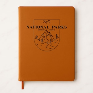 May include: A brown leather-bound journal with the words "NATIONAL PARKS" and a mountain illustration. The journal has a ribbon bookmark and is designed for memory keeping and journaling.