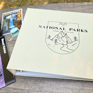 Może przedstawiać: Kremowy segregator z napisem "NATIONAL PARKS COLLECTION" i grafiką gór. Obok segregatora znajdują się lornetka, pocztówka z wodospadem i broszura o górze Rainier.