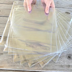 Può includere: Una pila di proteggi fogli di plastica trasparente, tenuti in mano, a ventaglio su una superficie di legno stagionato. Ogni foglio ha un bordo rinforzato con un foro per la rilegatura. I fogli sono rettangolari.